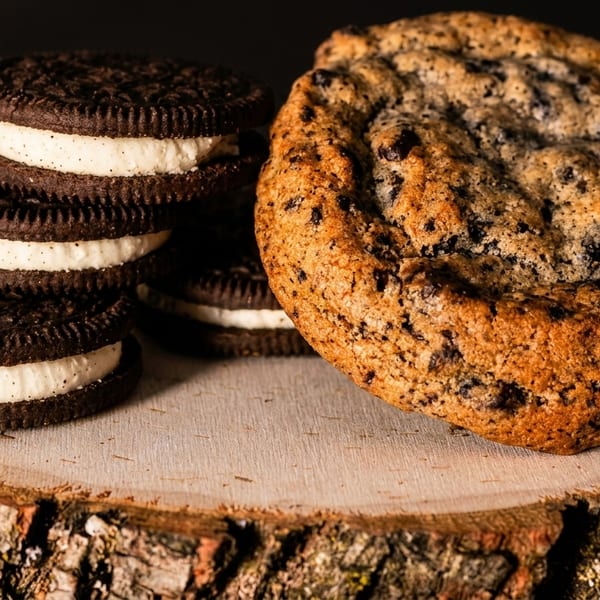 A stack of cream-filled chocolate sandwich cookies next to a large chocolate chip cookie on a wooden slab.